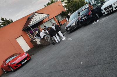 Des personnes discutent devant un bâtiment avec des voitures garées, dont une sportive rouge au premier plan.