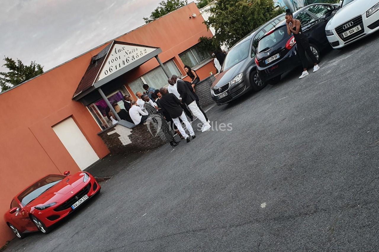 Des personnes discutent devant un bâtiment avec des voitures garées, dont une sportive rouge au premier plan.