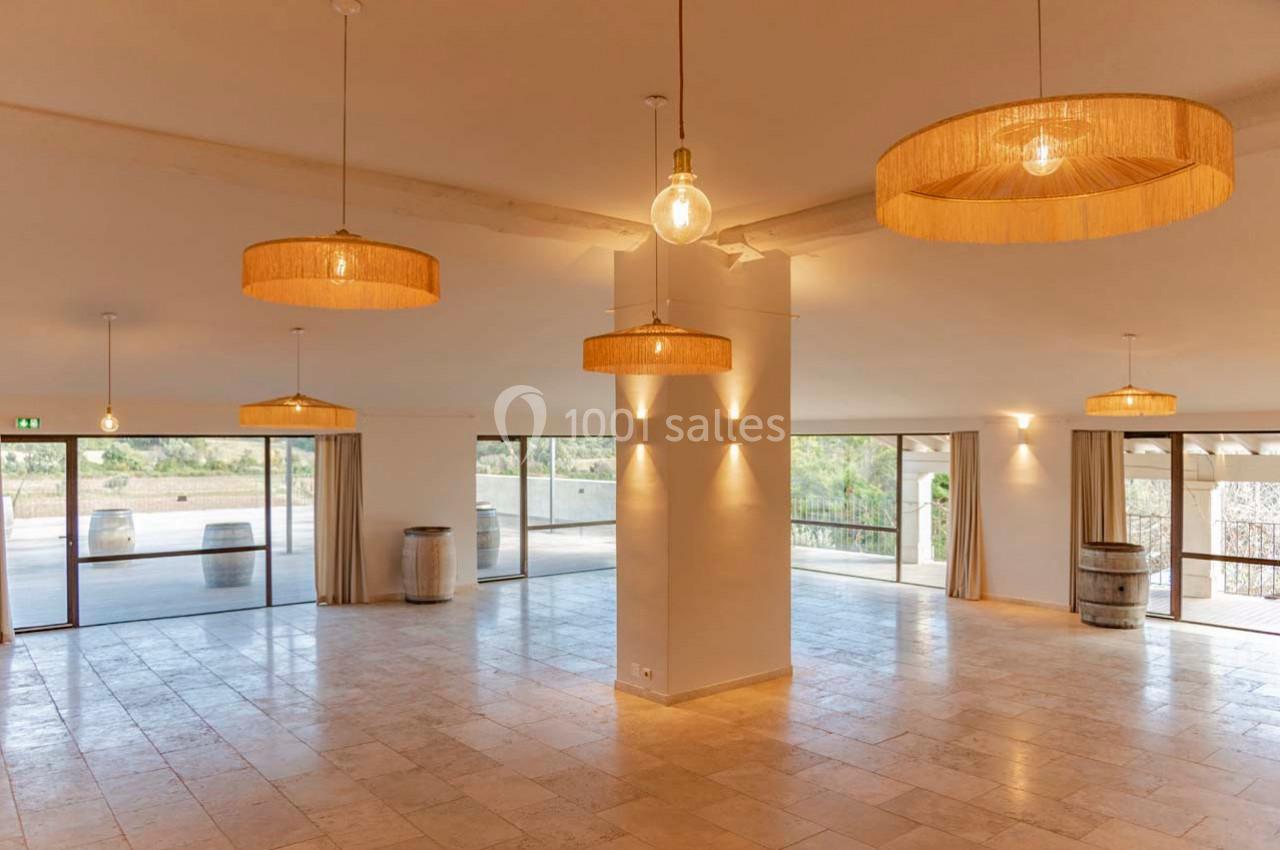 Salle lumineuse avec grandes baies vitrées, sol en pierre claire, suspensions en rotin et tonneaux décoratifs.