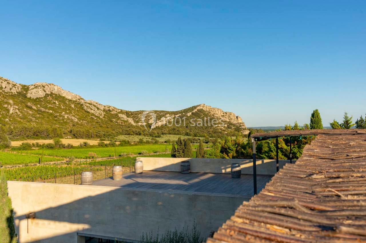 Vue d'une terrasse en bois avec des barriques, entourée de vignes et de collines sous un ciel dégagé.