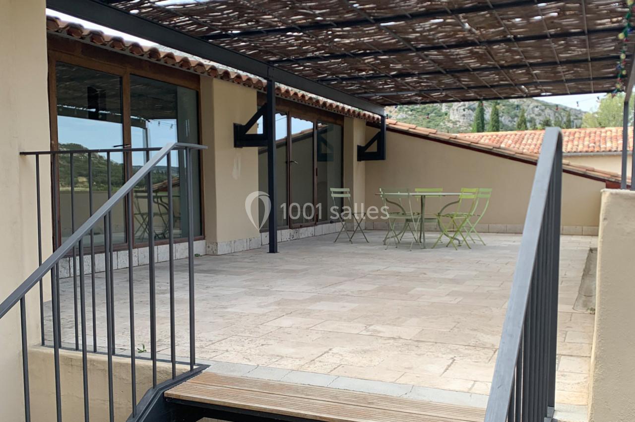 Terrasse en pierre avec table et chaises vertes sous une pergola en bois, entourée de bâtiments aux toits en tuiles.