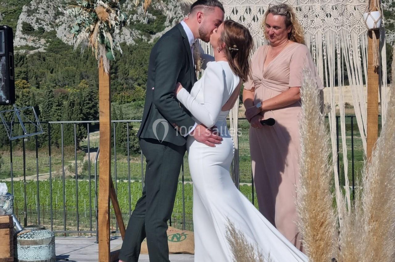 Un couple s'embrasse lors d'une cérémonie de mariage en extérieur, devant un décor naturel et boisé.