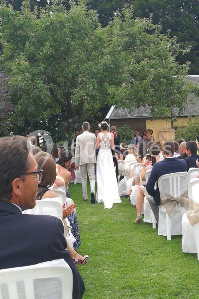 Un couple marche dans une allée extérieure lors d'une cérémonie de mariage, entouré d'invités assis.