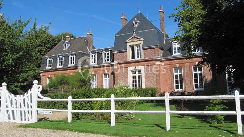 Façade d'une grande maison en briques rouges avec toit en ardoise, entourée de verdure et clôturée par une barrière blanche.
