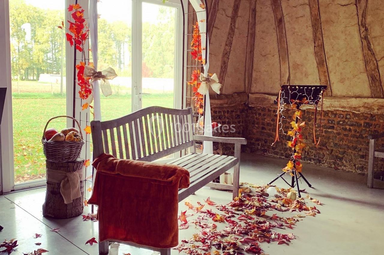 Banc en bois sous une arche décorée de feuilles d'automne, entouré de bougies et de feuilles au sol dans une salle rustique.