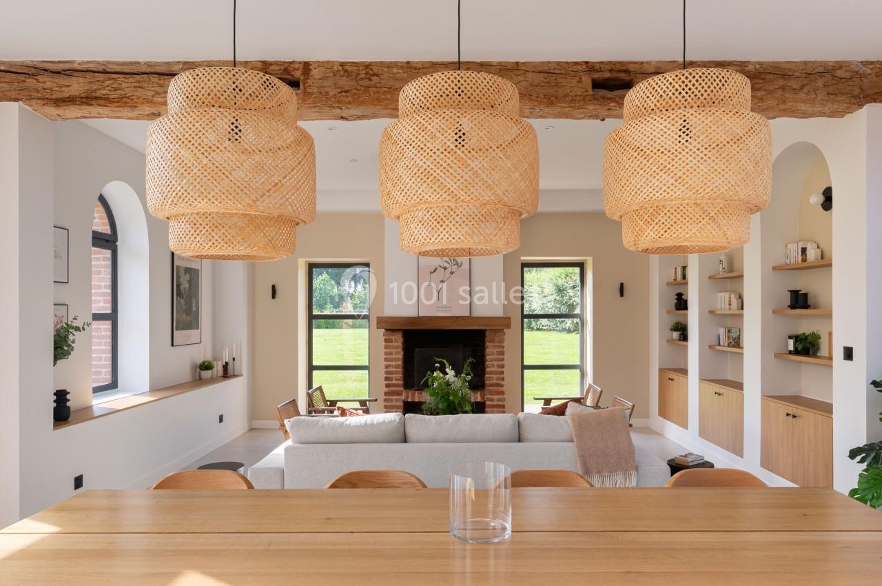 Salle à manger lumineuse avec table en bois, chaises assorties, suspensions en rotin et vue sur un salon avec cheminée.