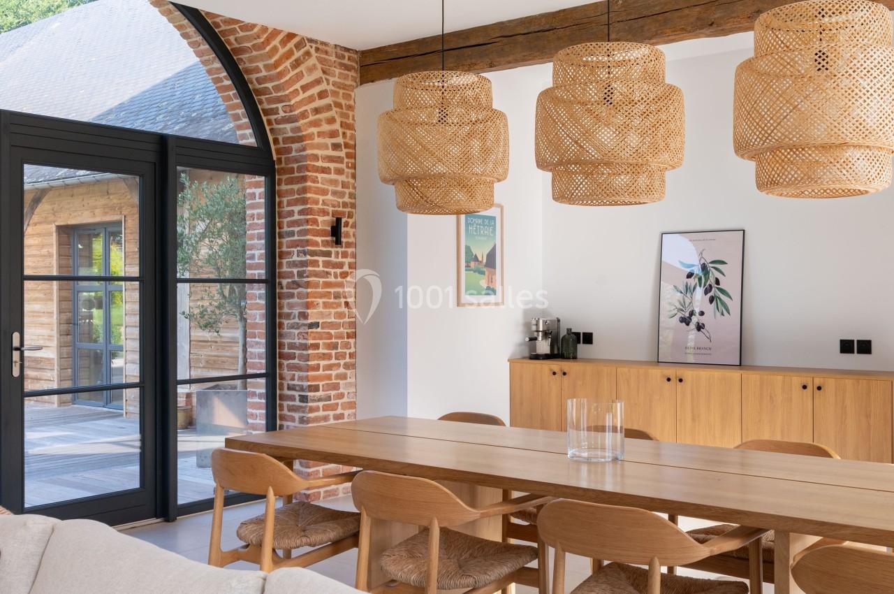 Salle à manger lumineuse avec table en bois, chaises assorties, suspensions en rotin et mur en briques apparentes.