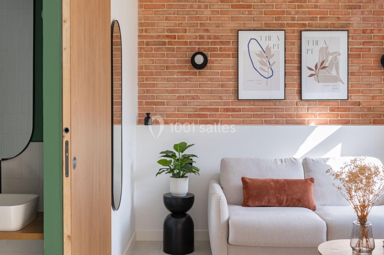 Salon lumineux avec mur en briques, canapé beige, coussin orange, plantes décoratives et affiches encadrées.