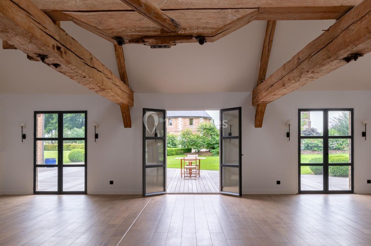 Vue intérieure d'une pièce lumineuse avec poutres apparentes, parquet, portes vitrées ouvertes sur un jardin.