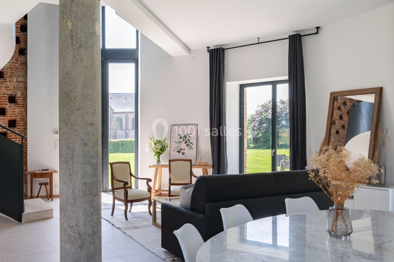 Salon lumineux avec mobilier moderne, table en marbre, canapé noir, grandes fenêtres et vue sur un jardin verdoyant.