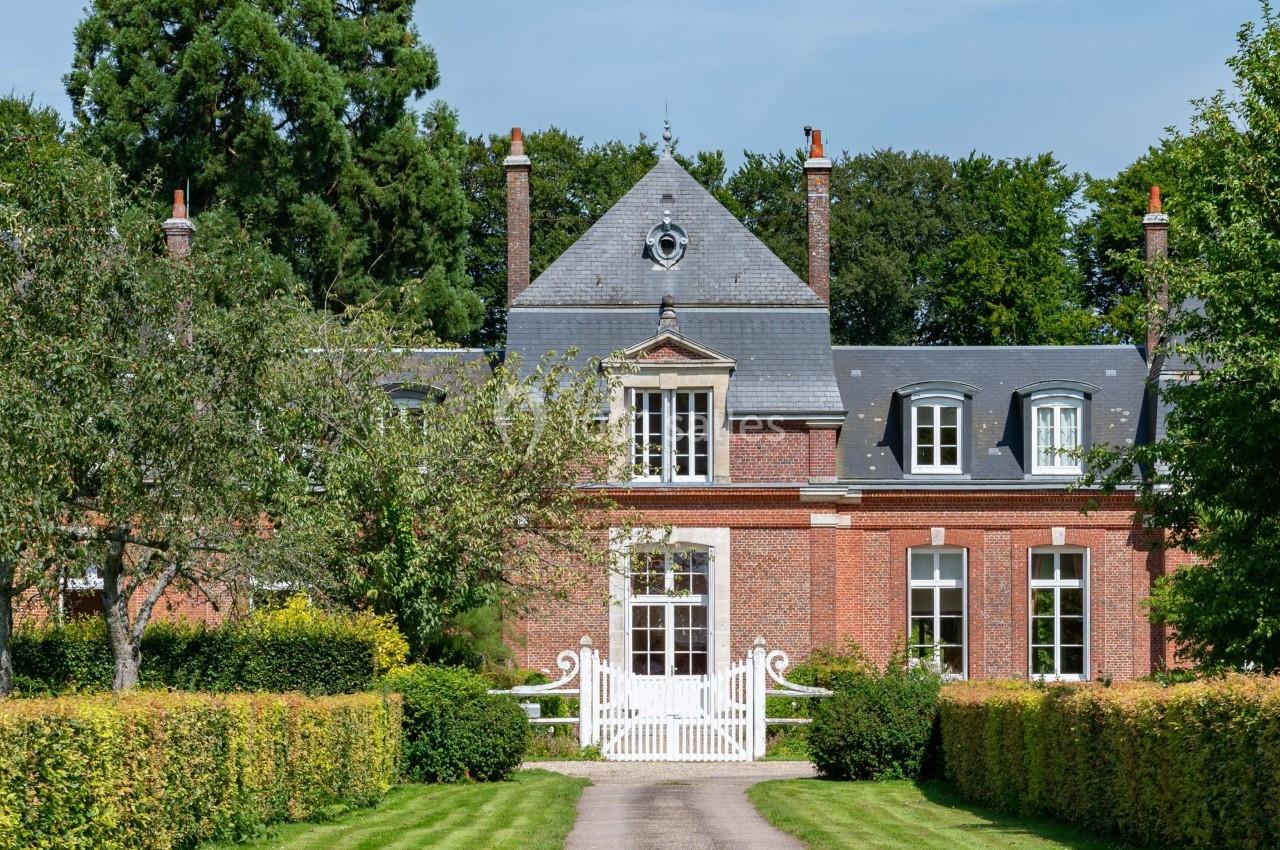 Allée bordée de haies menant à une maison en briques rouges avec toit en ardoise, entourée d'arbres.