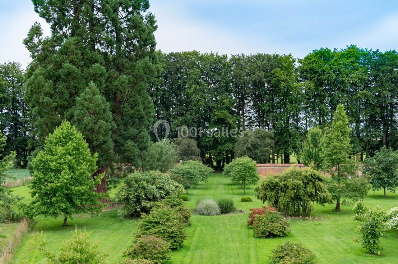 Vue d'un parc verdoyant avec pelouse, arbustes alignés, grands arbres et une haie en arrière-plan.
