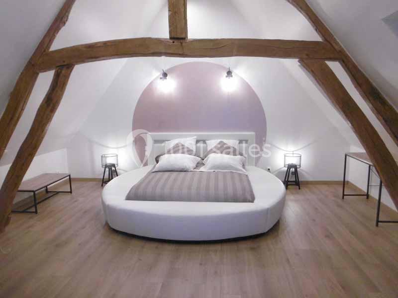 Chambre mansardée avec lit rond, poutres apparentes, éclairage mural et mobilier minimaliste.