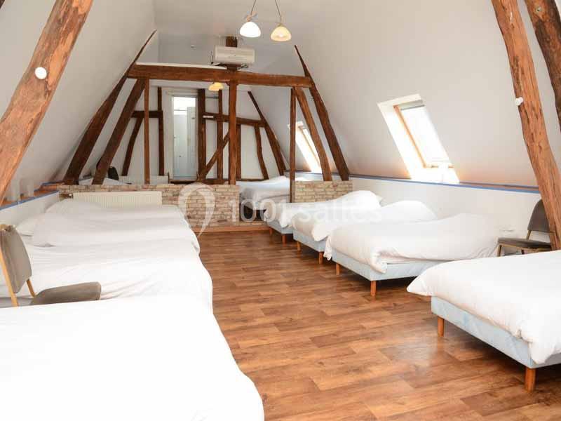 Dortoir mansardé avec plusieurs lits simples alignés, poutres apparentes et parquet en bois clair.