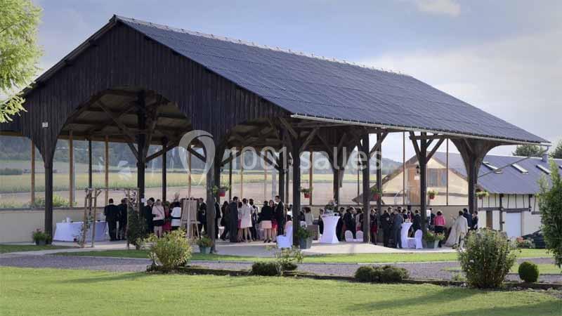 Structure en bois ouverte accueillant un événement avec des invités debout et des tables dressées, entourée de verdure.
