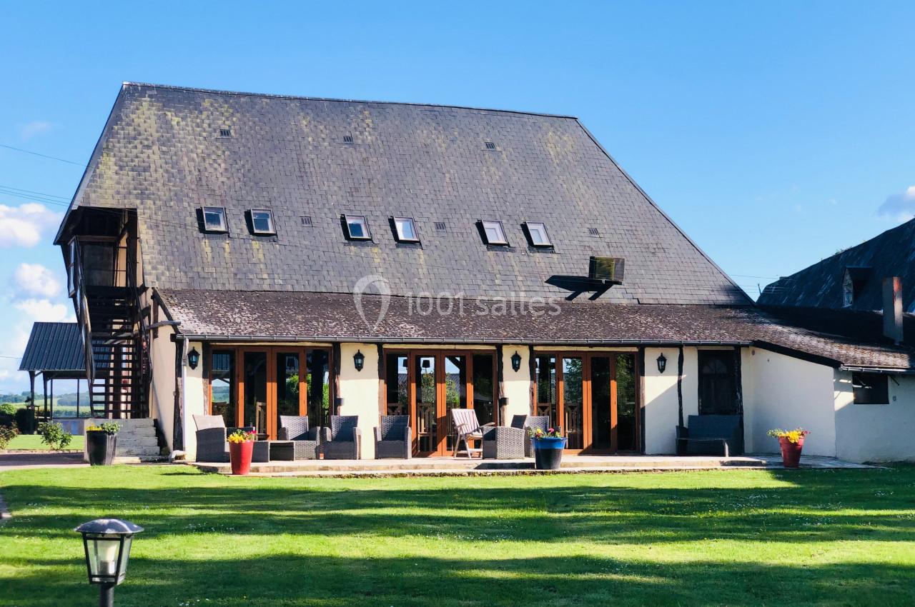 Grande maison avec toit en ardoise, nombreuses fenêtres et terrasse aménagée, entourée d'une pelouse verte.