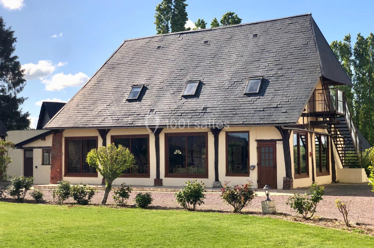 Maison à colombages avec toit en ardoise, fenêtres mansardées et jardin arboré sous un ciel dégagé.