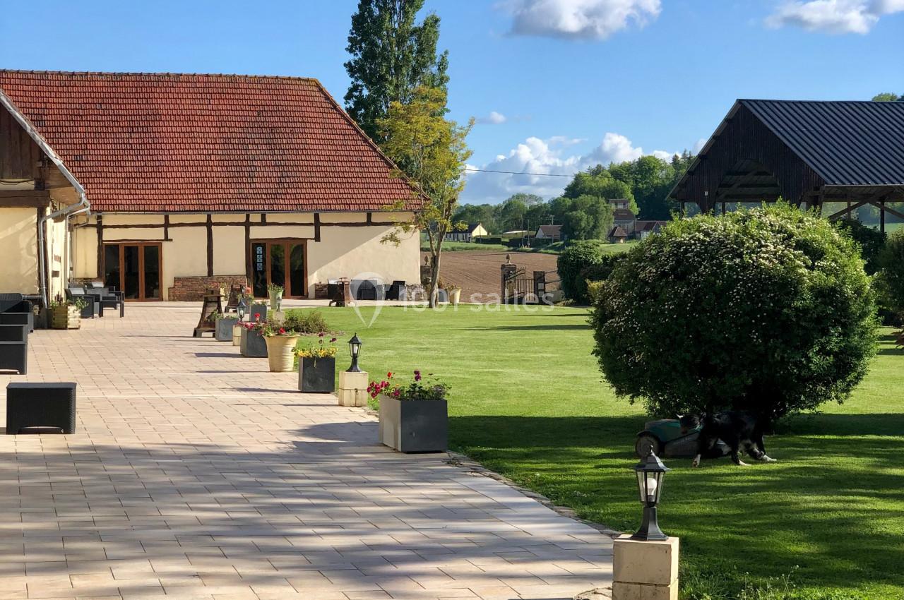 Allée pavée bordée de fleurs menant à des bâtiments en pierre et à une pelouse arborée sous un ciel dégagé.