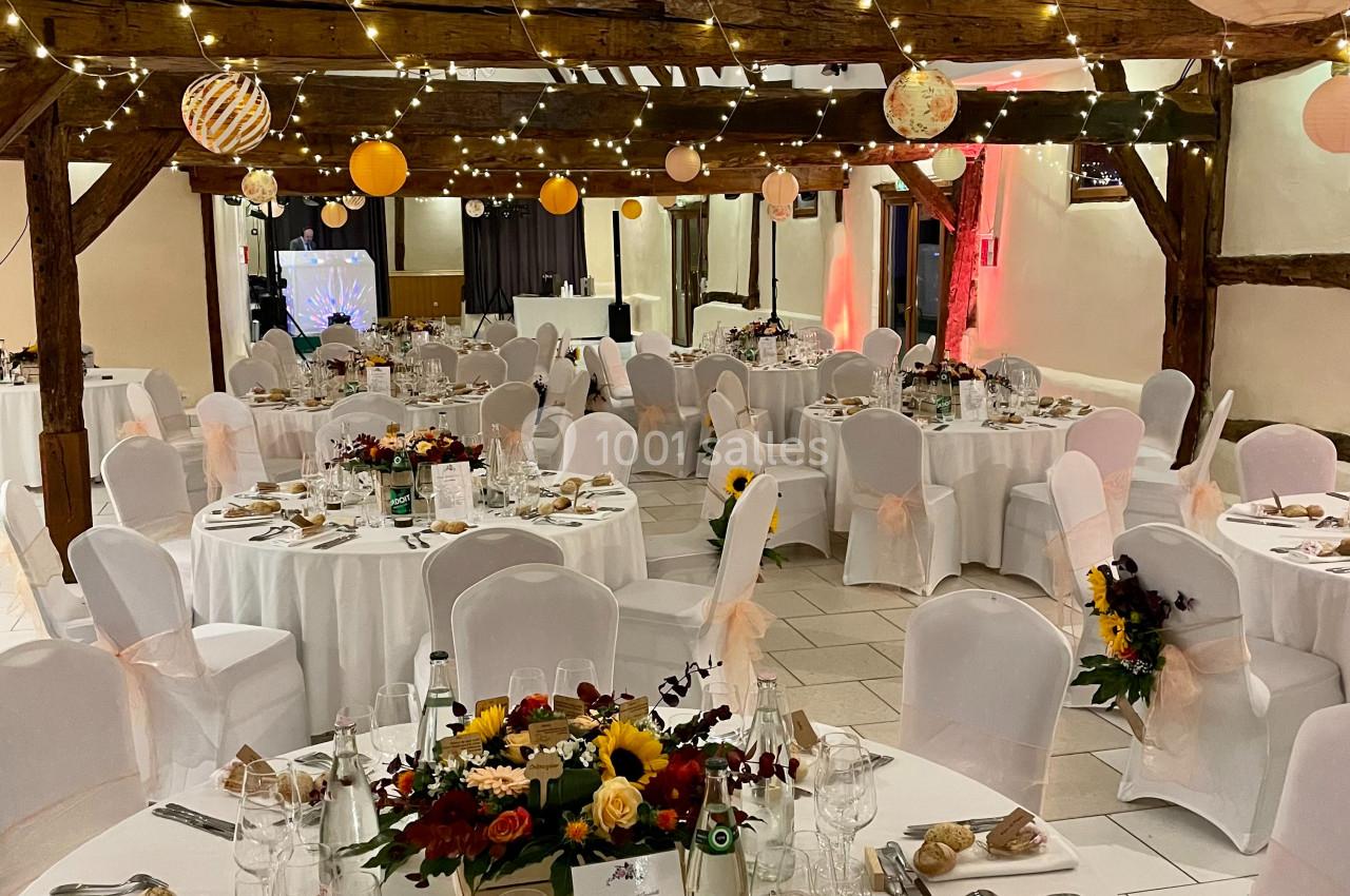 Salle de réception décorée avec des tables rondes dressées, des guirlandes lumineuses et des fleurs au centre des tables.