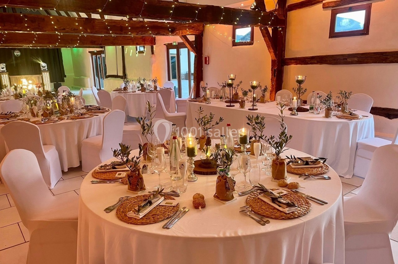 Salle de réception décorée avec des tables rondes, nappes blanches, bougies et éléments végétaux dans une ambiance…