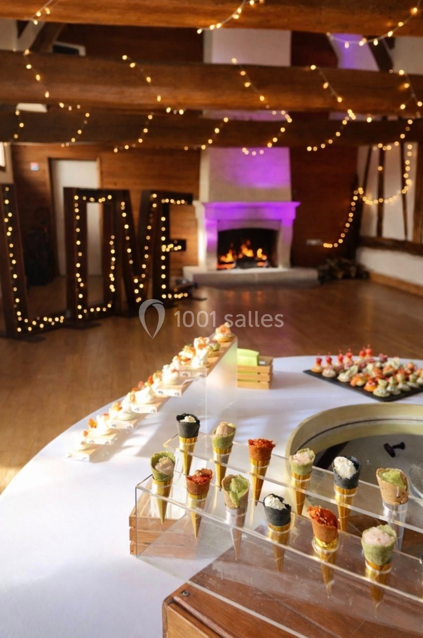 Buffet de réception avec cônes apéritifs, cheminée allumée et décor lumineux formant le mot ’LOVE’ en arrière-plan.