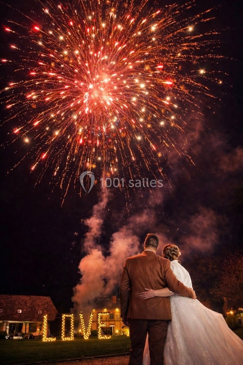 Un couple observe un feu d'artifice éclatant dans le ciel nocturne, avec le mot ’LOVE’ illuminé en arrière-plan.