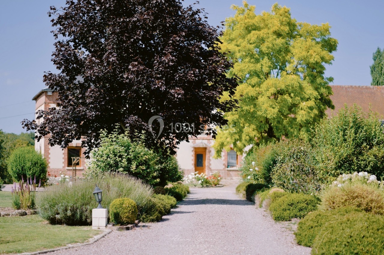 Allée gravillonnée bordée de buissons et d'arbustes menant à une maison entourée d'arbres.