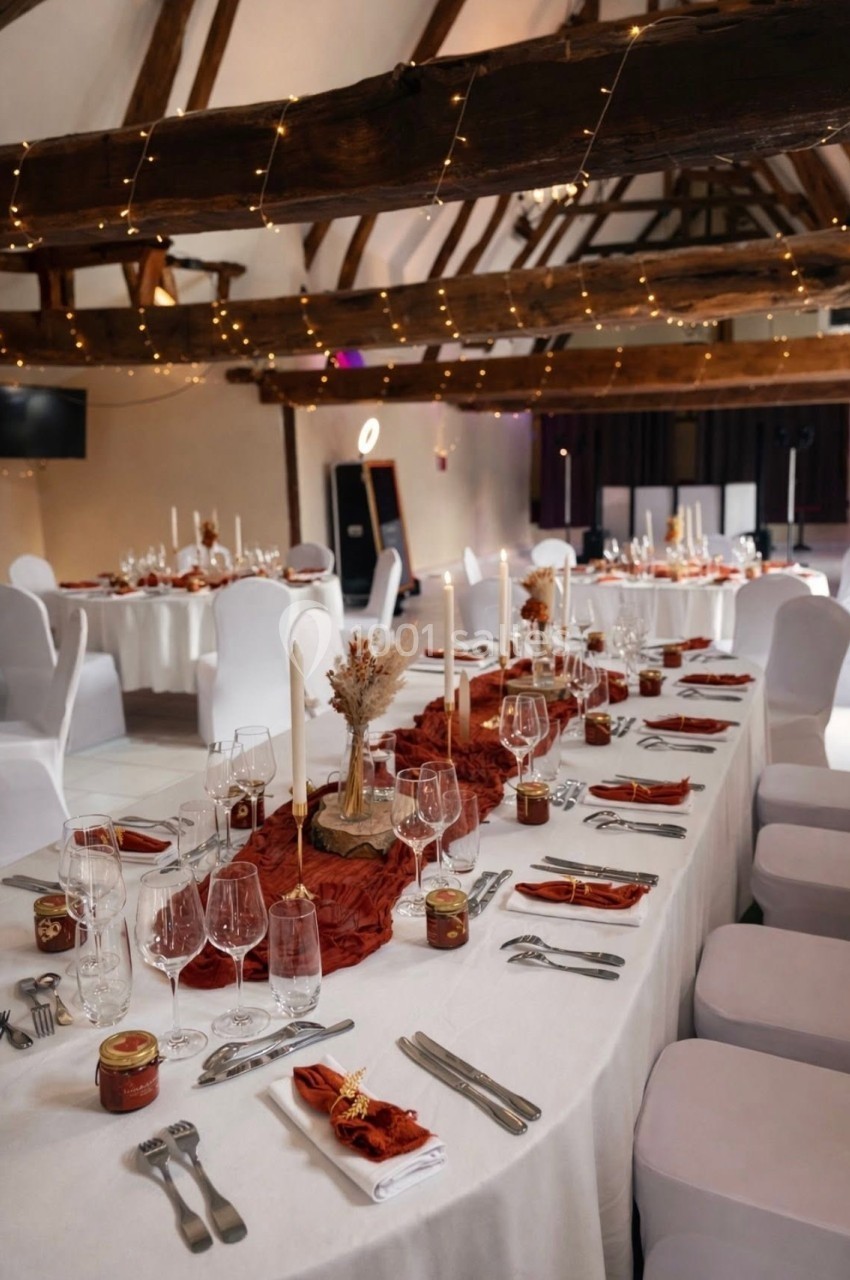 Salle de réception décorée avec des tables dressées, nappes blanches, bougies et accents rouges sous une charpente en bois.