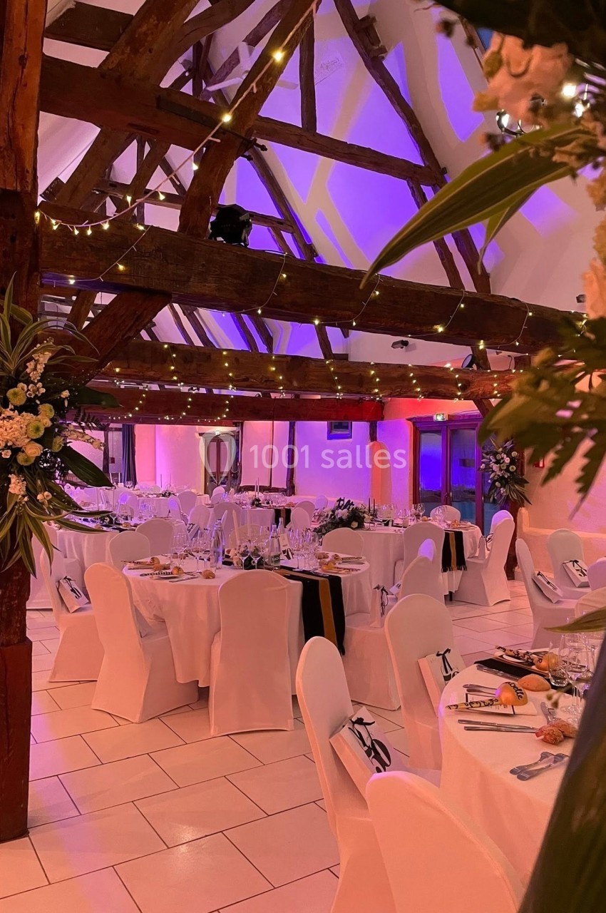Salle de réception décorée avec des tables dressées, des chaises blanches et des guirlandes lumineuses sous une charpente en…