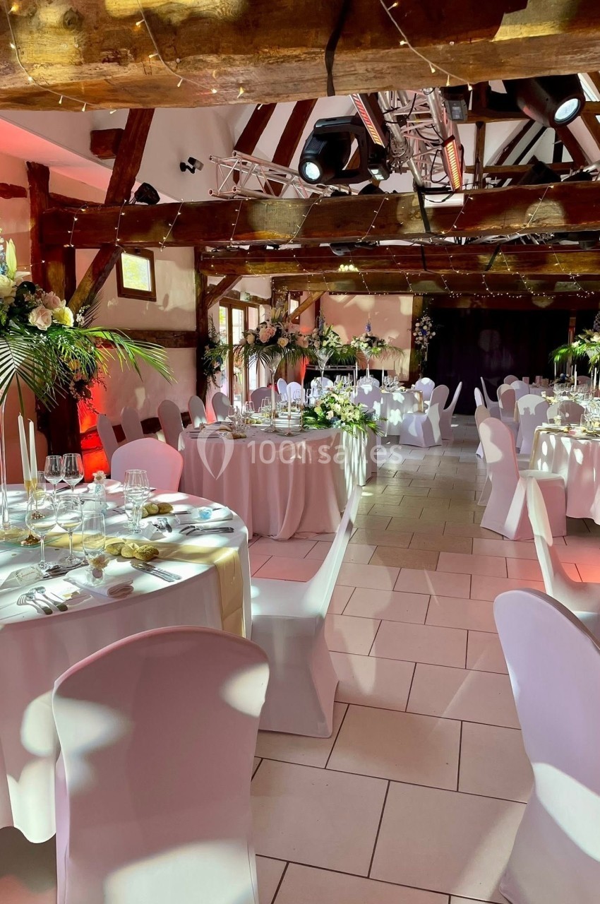 Salle de réception décorée avec des tables rondes, nappes blanches, fleurs et éclairage chaleureux sous des poutres en bois.