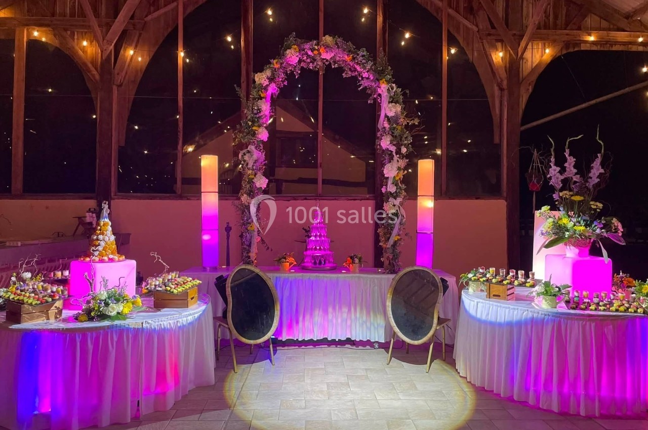 Salle décorée pour un événement avec arche florale, tables garnies et éclairage rose et chaleureux.