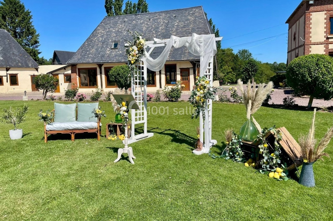 Décoration de mariage en extérieur avec arche fleurie, banc, et éléments végétaux sur une pelouse devant des bâtiments.