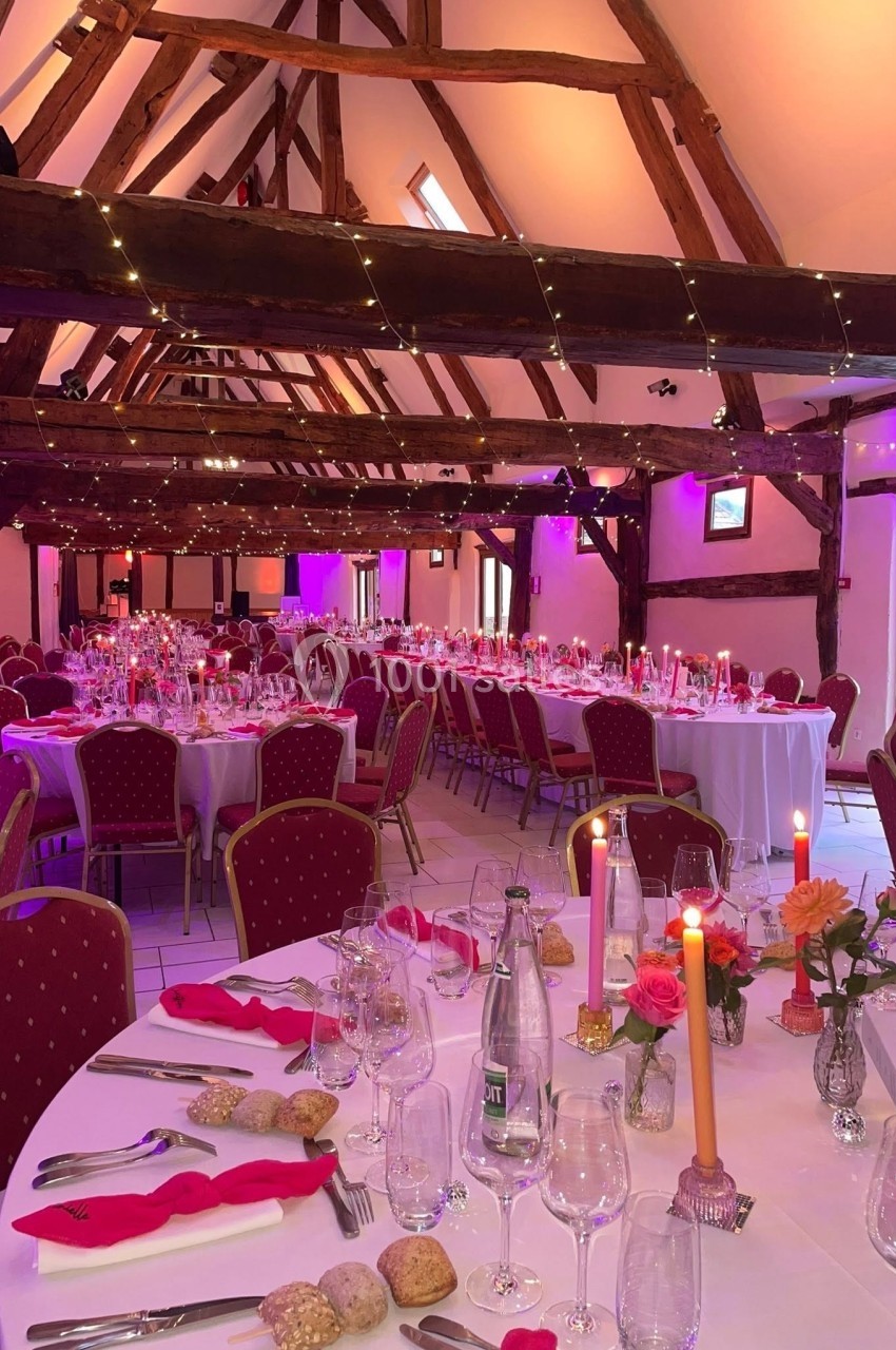 Salle de réception décorée avec des tables dressées, bougies allumées et éclairage d'ambiance rose sous une charpente en…
