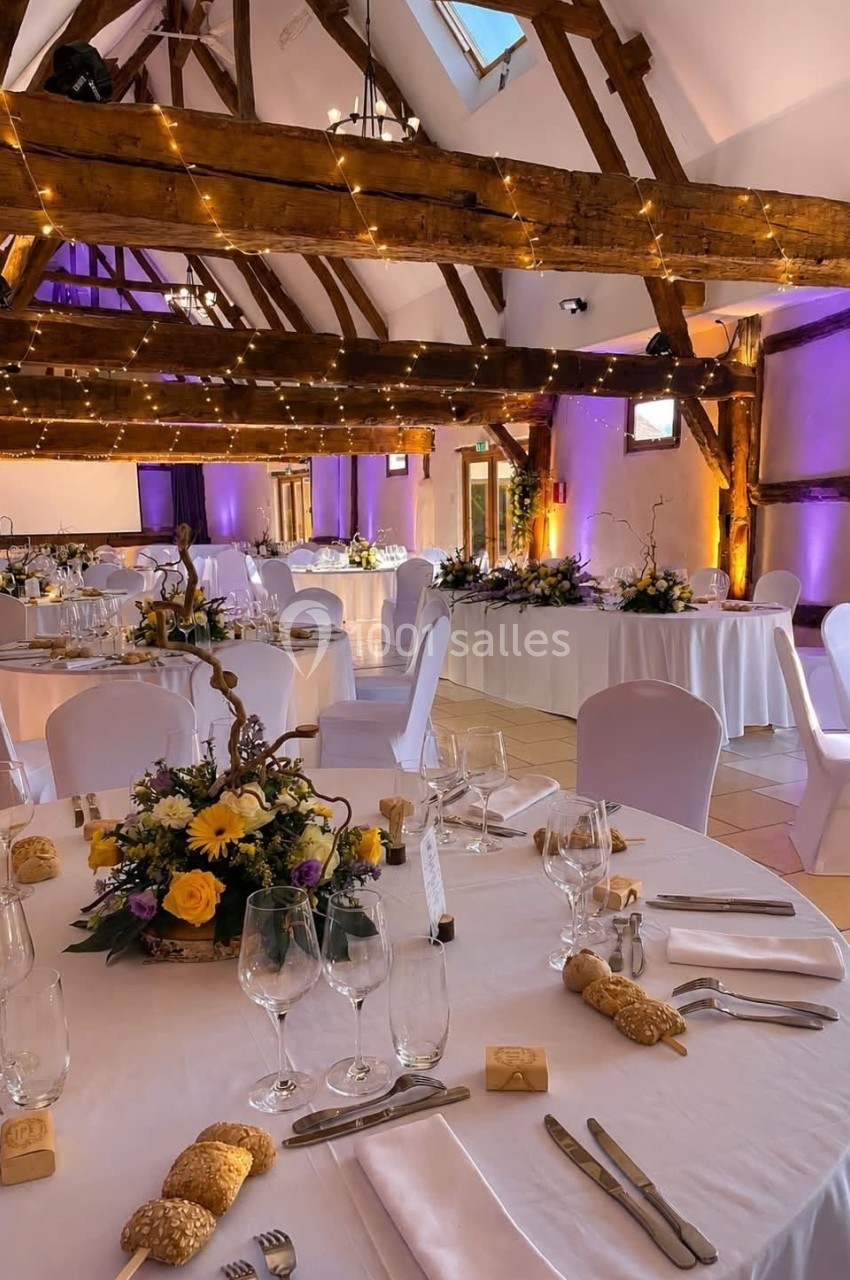 Salle de réception décorée avec des tables rondes dressées, des fleurs et des guirlandes lumineuses suspendues.