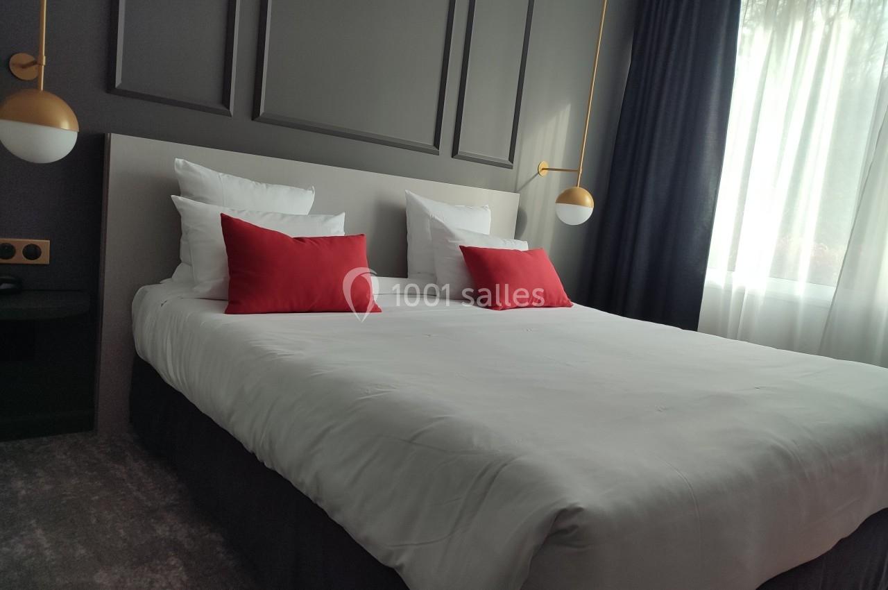 Lit double avec draps blancs, coussins rouges, suspensions dorées et lumière naturelle entrant par une grande fenêtre.