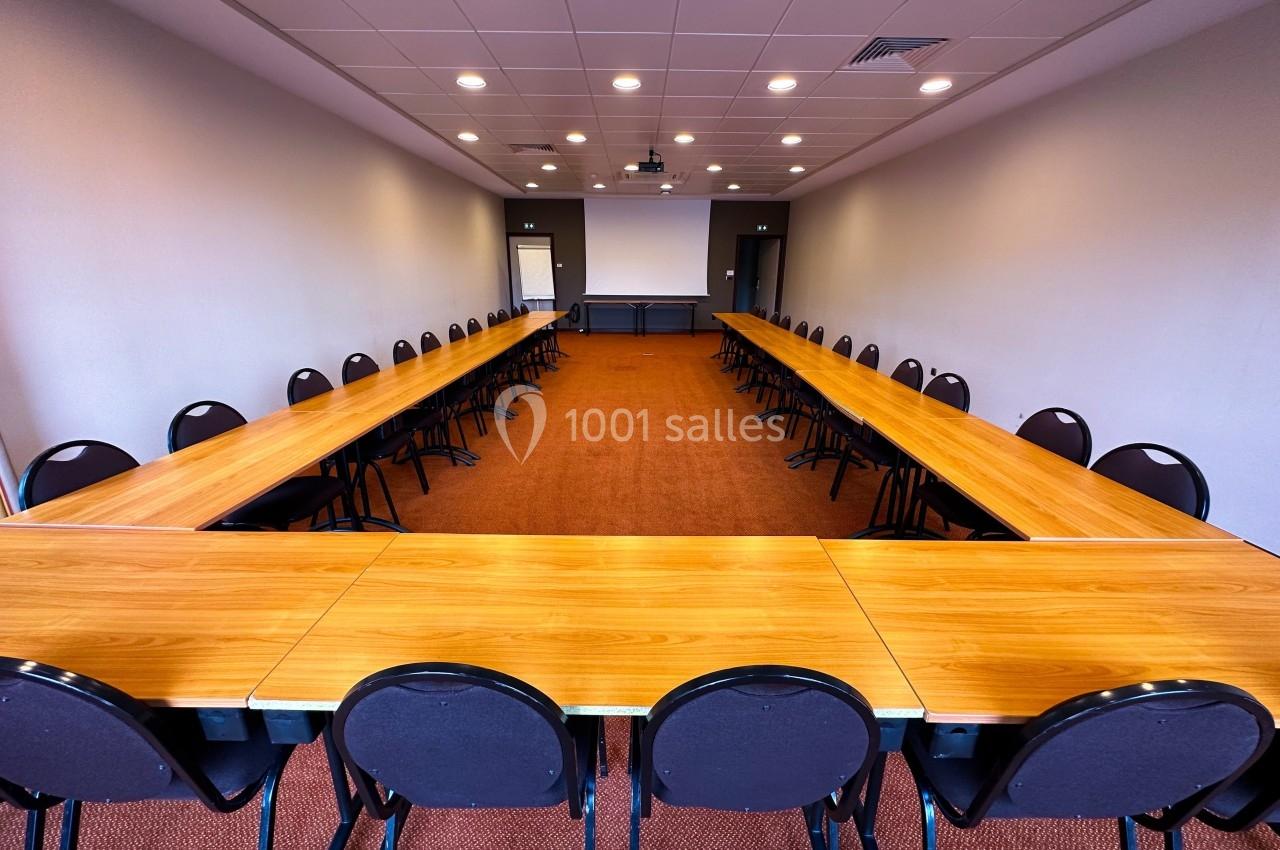 Salle de réunion avec des tables disposées en U, entourées de chaises noires, éclairée par des plafonniers.