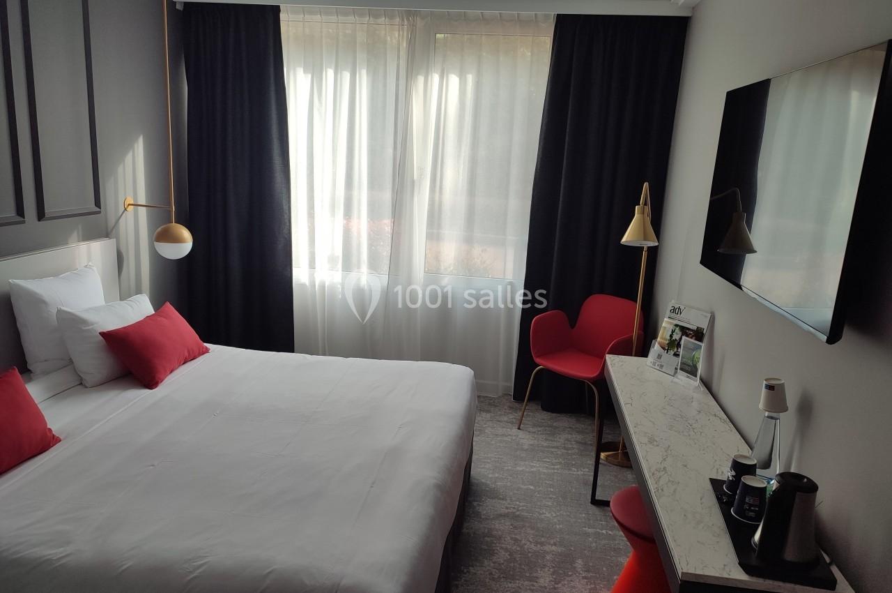 Chambre d'hôtel lumineuse avec un lit double, des coussins rouges, un bureau, une chaise et une télévision murale.