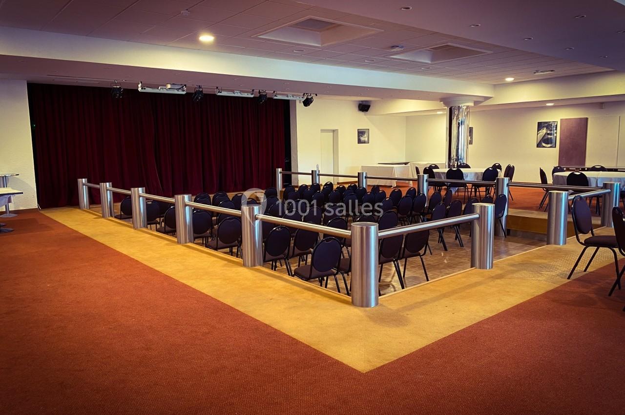 Salle de conférence vide avec des rangées de chaises noires, une scène avec rideaux rouges et un sol en moquette.
