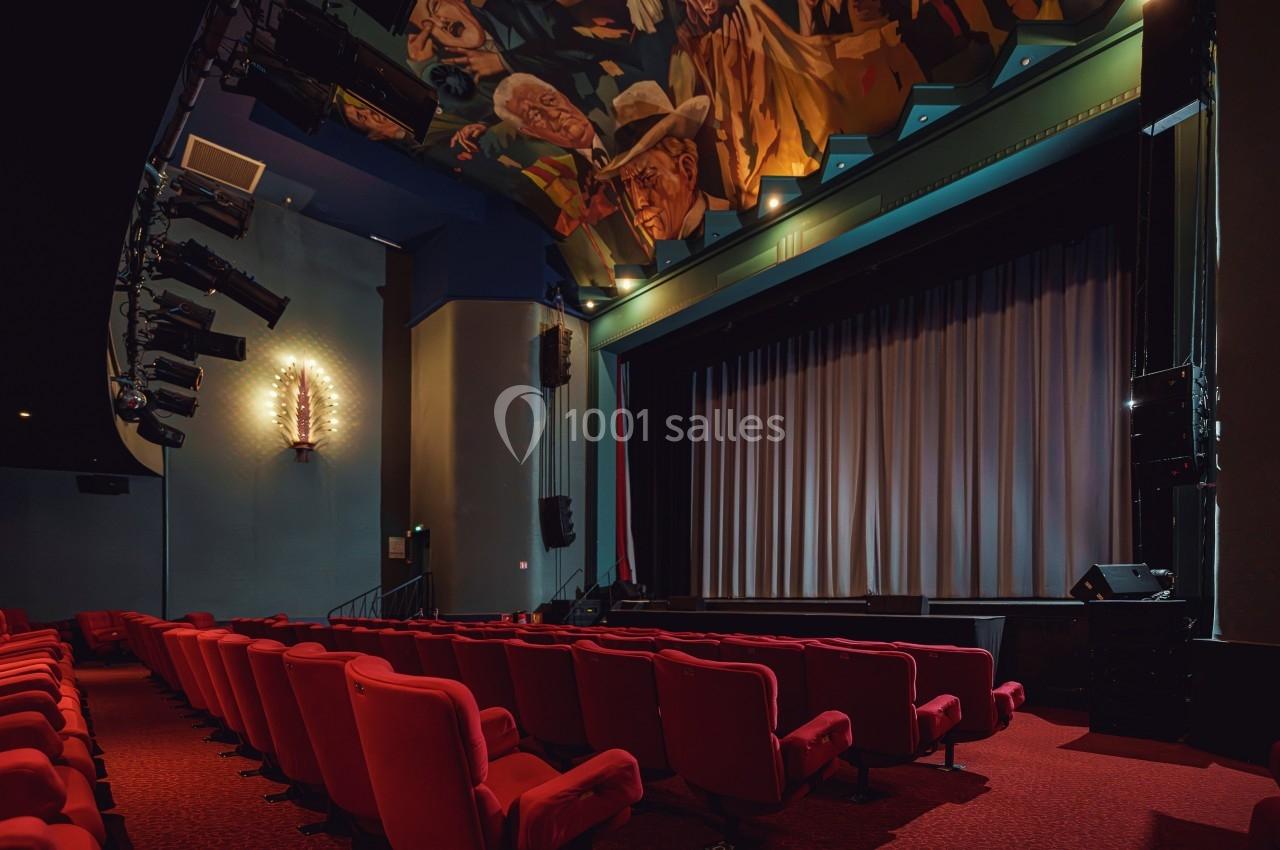 Salle de théâtre vide avec des fauteuils rouges, une scène avec rideau fermé et un plafond décoré de fresques.
