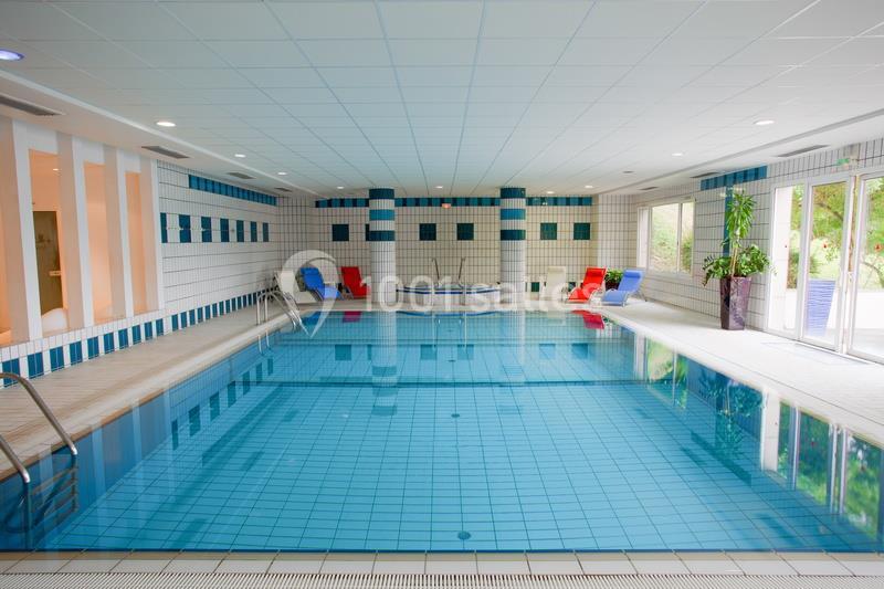 Piscine intérieure avec carrelage bleu, transats colorés et grandes baies vitrées laissant entrer la lumière naturelle.