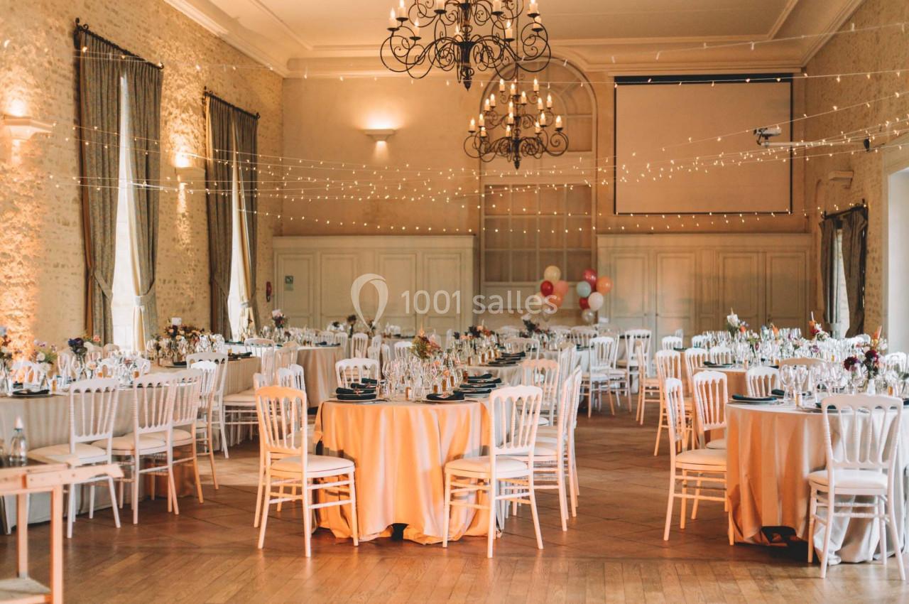 Salle de réception élégamment décorée avec des tables rondes, des chaises blanches et des guirlandes lumineuses suspendues.
