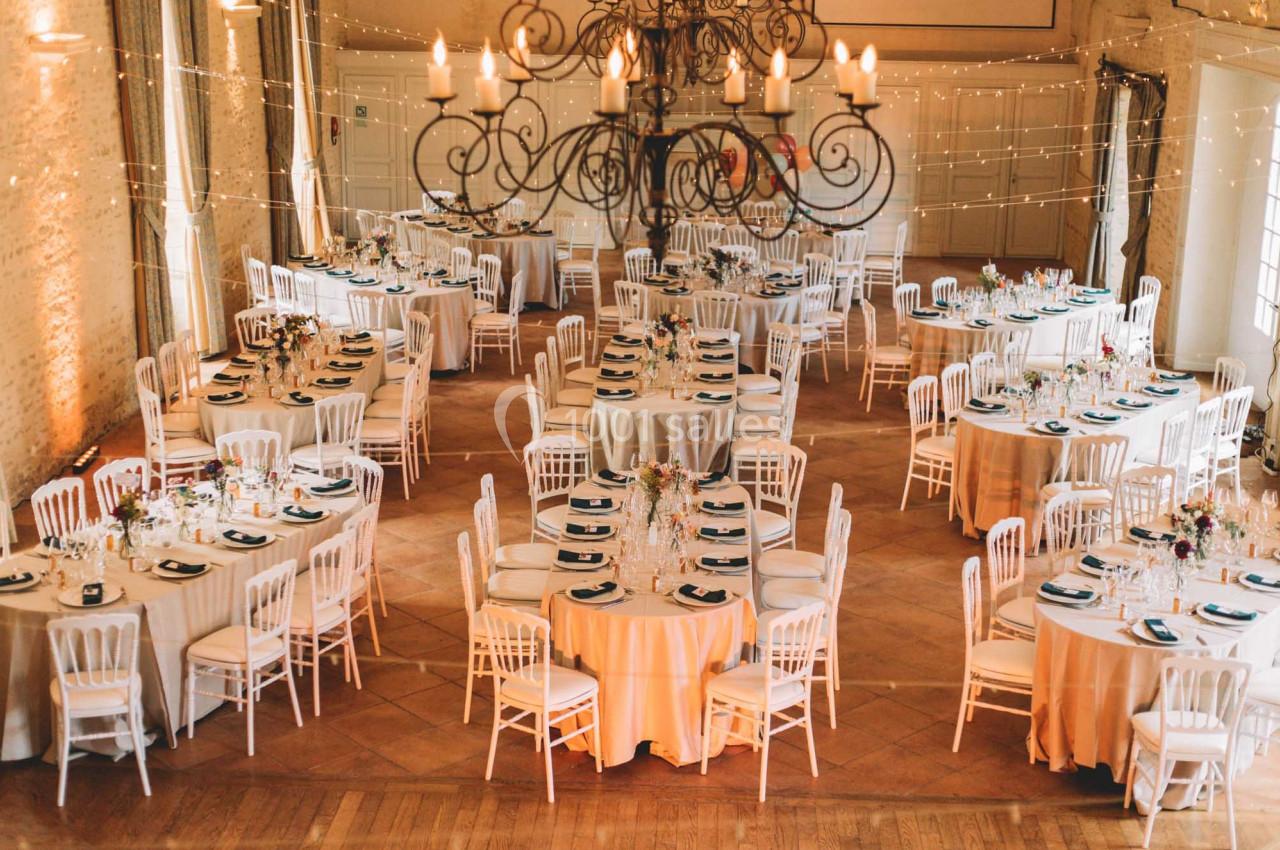 Salle de réception décorée avec des tables rondes dressées, des chaises blanches et un grand lustre central.