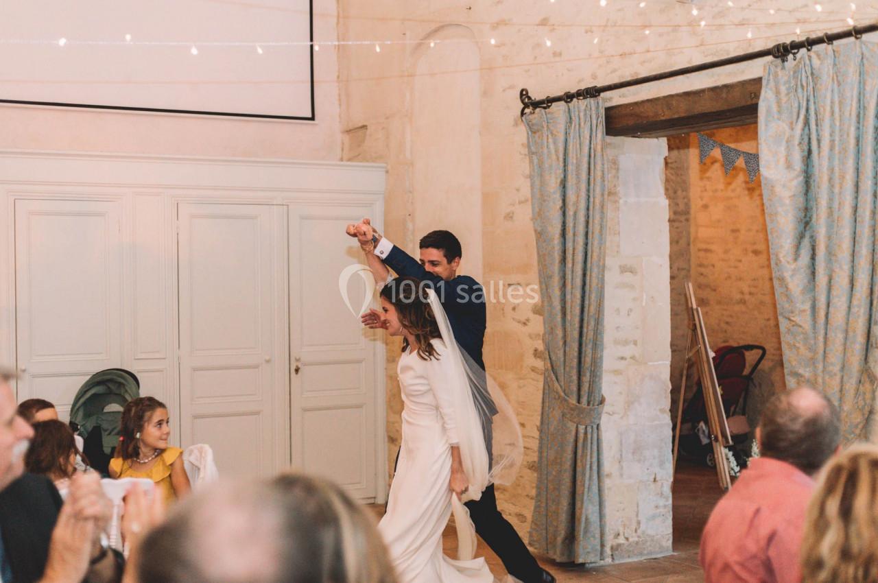 Un couple de mariés danse dans une salle décorée, sous des guirlandes lumineuses, entouré d'invités assis.
