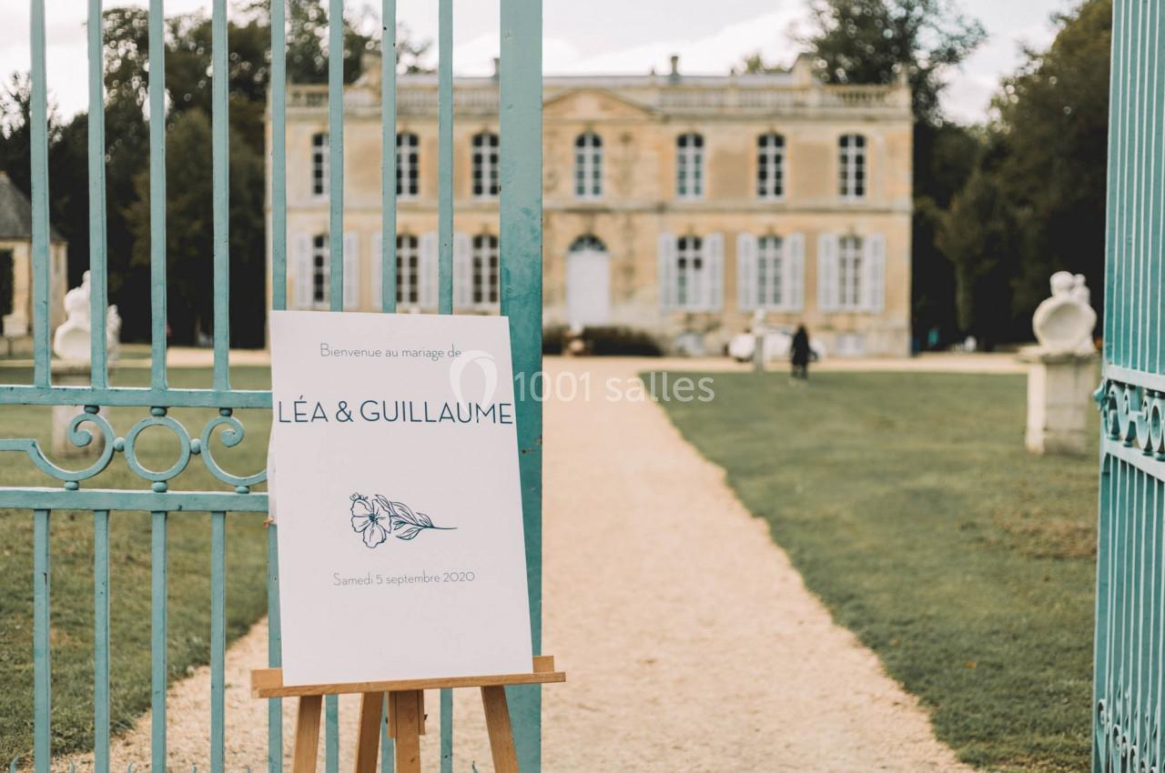 Panneau de bienvenue à un mariage devant un portail ouvert menant à un château entouré de verdure.