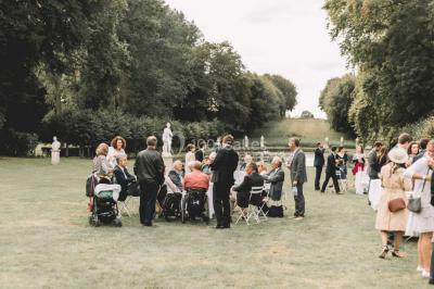Groupe de personnes rassemblées dans un jardin près d'une bâtisse ancienne, sous une tente blanche par temps clair.