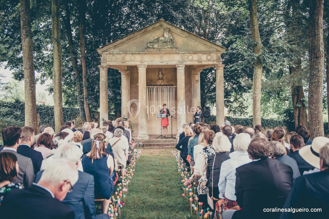Cérémonie en plein air devant un petit temple entouré d'arbres, avec des invités assis et une personne parlant au micro.