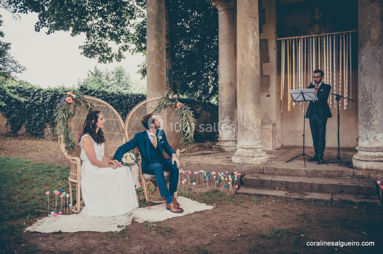 Un couple assis lors d'une cérémonie en plein air, écoutant un discours d'un homme debout avec un micro.