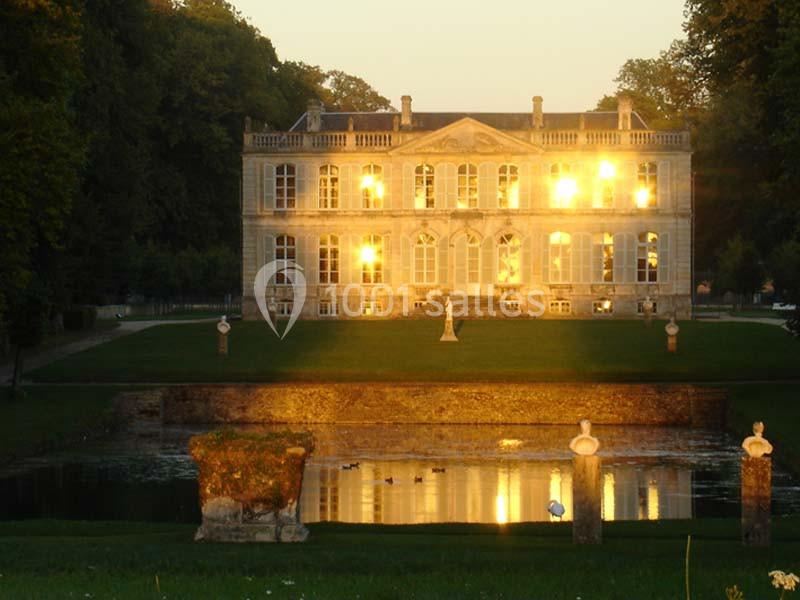 Façade d'un château éclairée par le soleil couchant, avec un jardin, un bassin et des statues au premier plan.