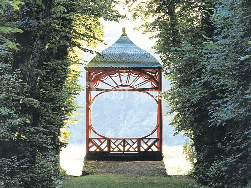 Kiosque en bois rouge avec toit en ardoise, entouré d'arbres et baigné de lumière naturelle.