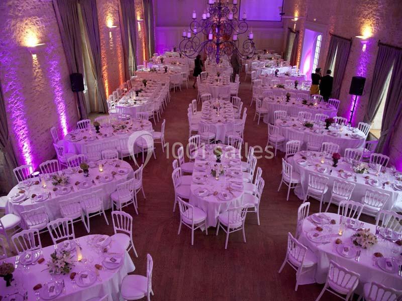 Salle de réception décorée avec des tables rondes, nappes blanches, chaises blanches et éclairage violet.